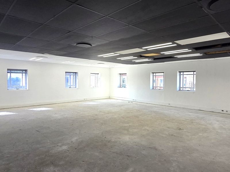 A large, open-plan office space featuring a suspended ceiling with recessed lighting, concrete flooring, and multiple windows providing natural light.