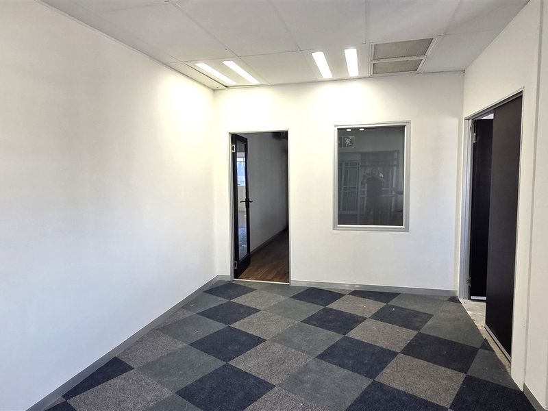 A clean, empty office space featuring neutral walls, checkered carpet tiles, recessed ceiling lighting, and an internal glass partition for natural light flow.