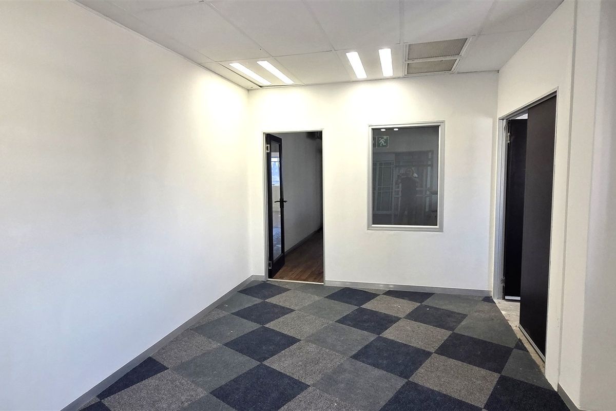 A clean, empty office space featuring neutral walls, checkered carpet tiles, recessed ceiling lighting, and an internal glass partition for natural light flow.