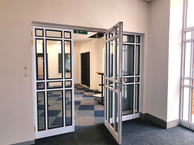 An empty office space featuring glass-paneled double doors, carpet tiles, and large windows, providing a bright and professional work environment.
