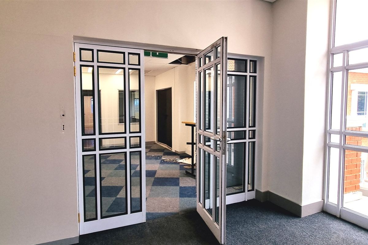 An empty office space featuring glass-paneled double doors, carpet tiles, and large windows, providing a bright and professional work environment.