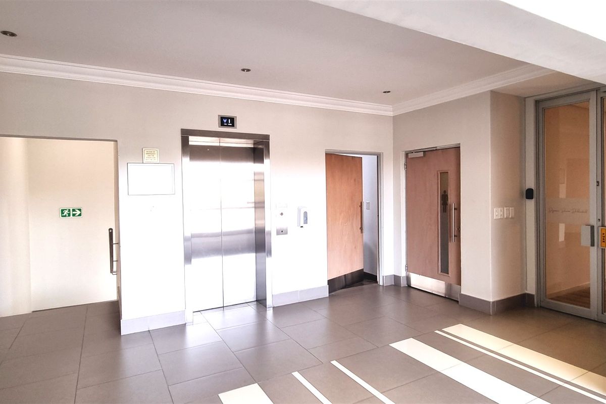 A clean and professional office building lobby featuring tiled flooring, a modern elevator, and glass-paneled entry doors with recessed lighting.