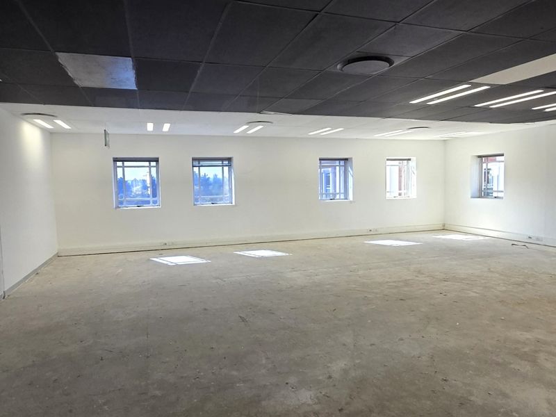 A spacious, vacant open-plan office area featuring a suspended ceiling, recessed lighting, and multiple windows providing natural light.