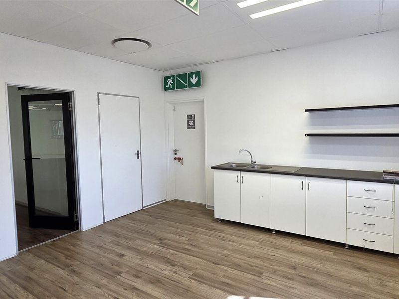 A clean, functional office kitchenette featuring white cabinetry, a stainless steel sink, laminate countertops, and floating shelves, set against a neutral interior.