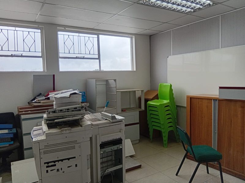 A functional office space featuring suspended ceiling tiles, large windows with security bars, and tiled flooring, suitable for a workspace or administrative area.