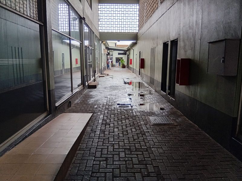 A covered outdoor walkway between office building wings, featuring paved brick flooring, glass storefronts, and decorative breeze block ventilation screens.