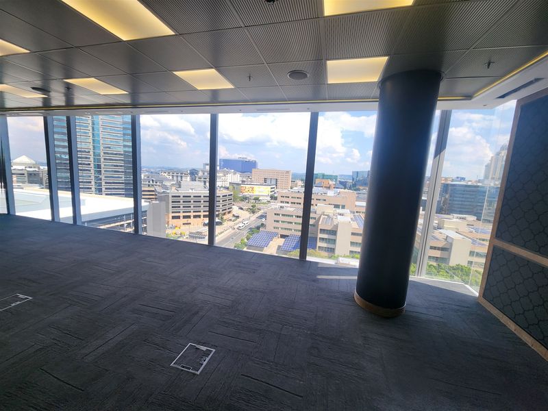 A spacious, empty office area featuring floor-to-ceiling windows with expansive city views, carpeted flooring, and modern recessed ceiling lighting.