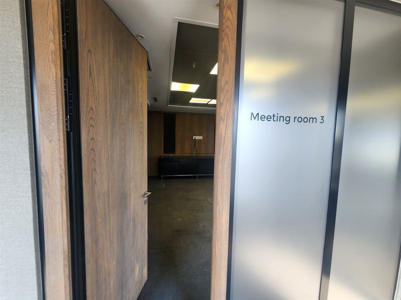 An empty office meeting room featuring wood paneling, carpet flooring, and a modern frosted glass entrance door.