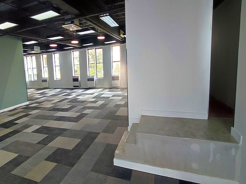 An open-plan office space featuring carpet tiles, exposed ceiling with recessed lighting, and multiple window air conditioning units.