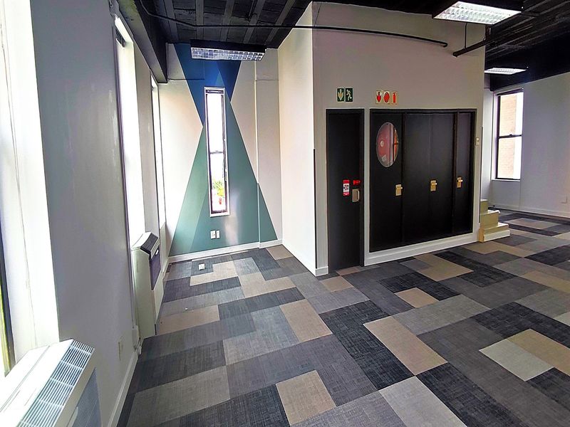 A bright, open-plan office space featuring modern carpet tiles, exposed industrial-style ceilings, and built-in storage cabinets.