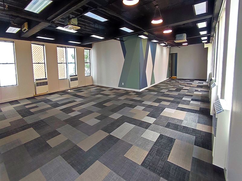 Spacious open-plan office area featuring exposed black ceilings, modern geometric wall accents, patterned carpet tiles, and multiple window-mounted air conditioning units.