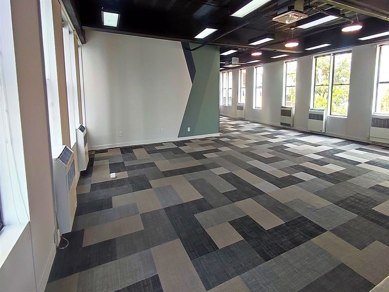 Spacious, open-plan office area featuring modern carpet tiles, large windows for natural light, and exposed ceiling with integrated lighting.