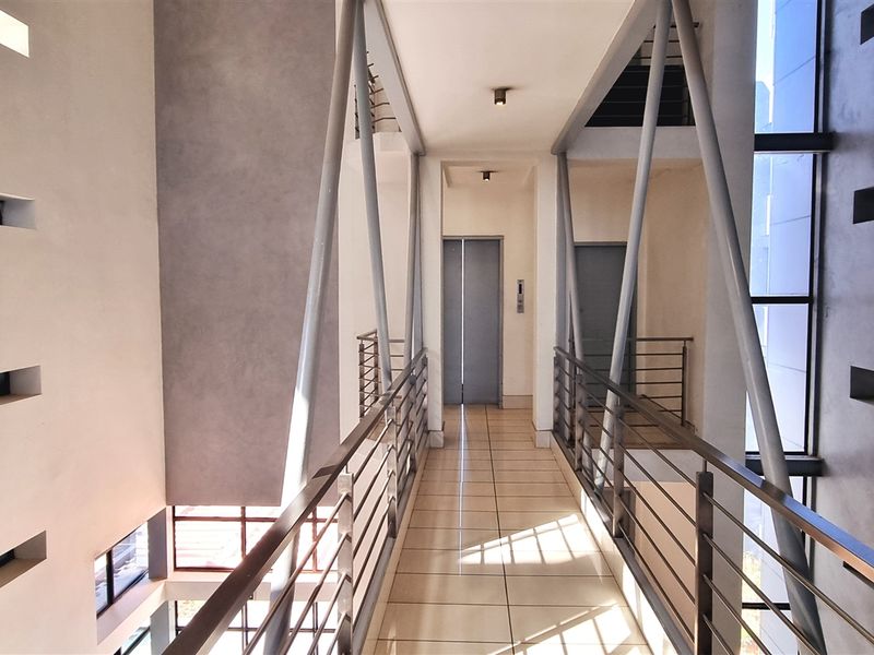 Modern office building interior featuring an elevated walkway with steel structural supports, tiled flooring, and elevator access.