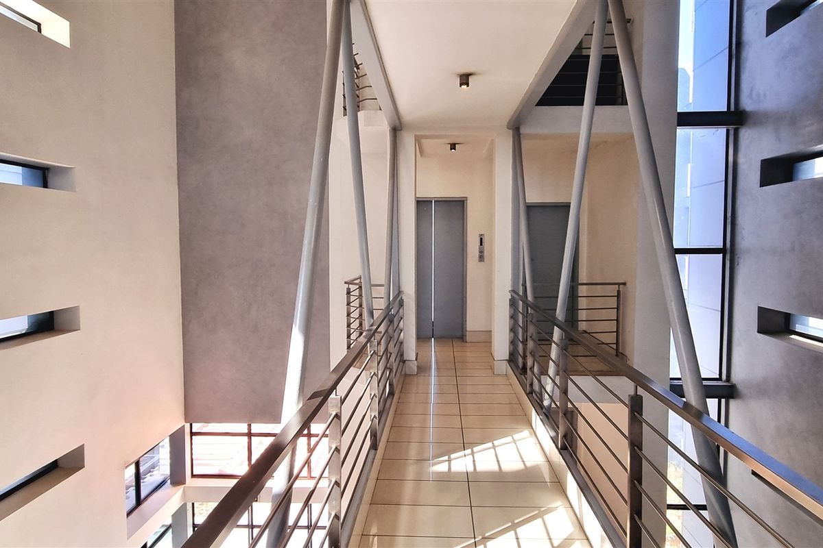 Modern office building interior featuring an elevated walkway with steel structural supports, tiled flooring, and elevator access.
