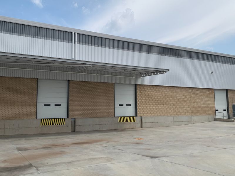 Industrial warehouse exterior featuring multiple loading dock roller shutter doors, concrete apron, and modern metal cladding with a protective canopy.
