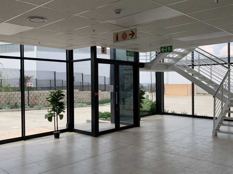 Modern industrial entrance lobby featuring floor-to-ceiling glass windows, tiled flooring, and a prominent metal staircase leading to the upper level.