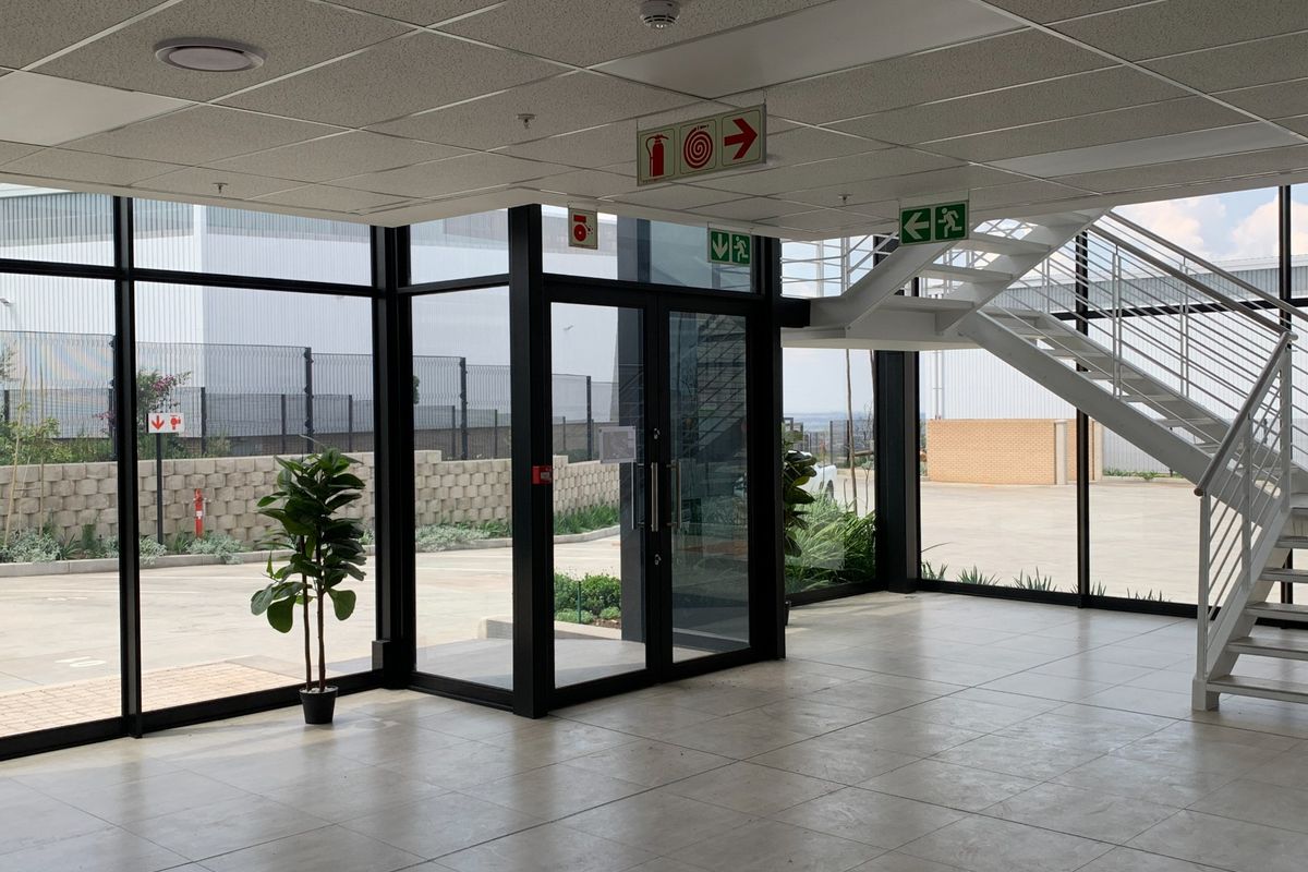 Modern industrial entrance lobby featuring floor-to-ceiling glass windows, tiled flooring, and a prominent metal staircase leading to the upper level.