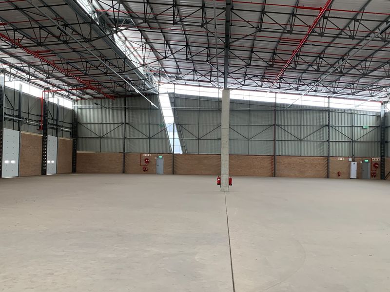 Spacious industrial warehouse featuring high ceilings with exposed steel trusses, concrete flooring, fire sprinkler system, and large roller shutter access doors.