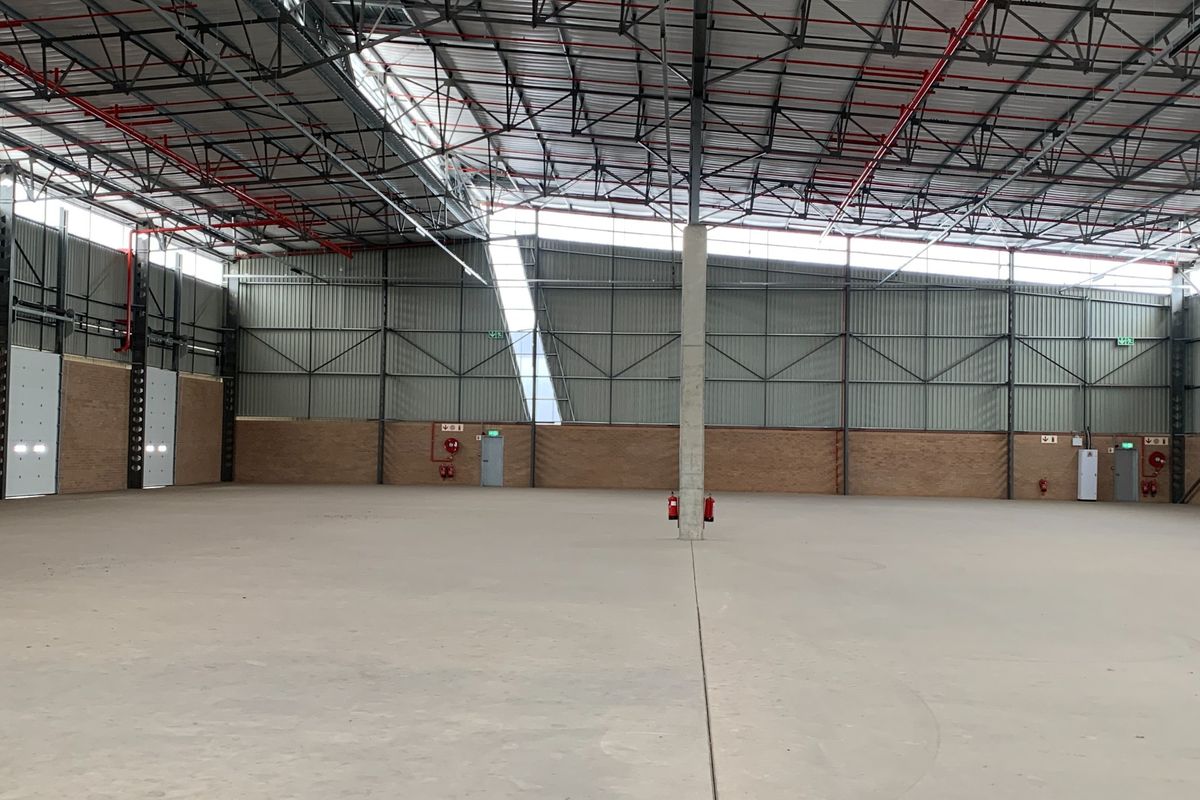 Spacious industrial warehouse featuring high ceilings with exposed steel trusses, concrete flooring, fire sprinkler system, and large roller shutter access doors.