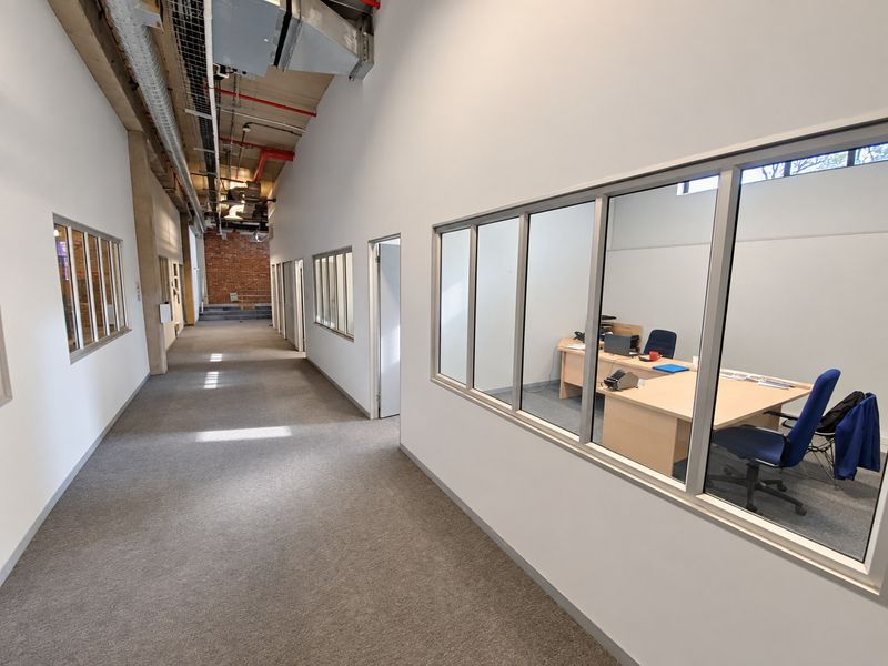 A modern industrial-style hallway featuring exposed ceiling services, carpeted flooring, and glass-partitioned office spaces with an exposed brick feature wall at the end.