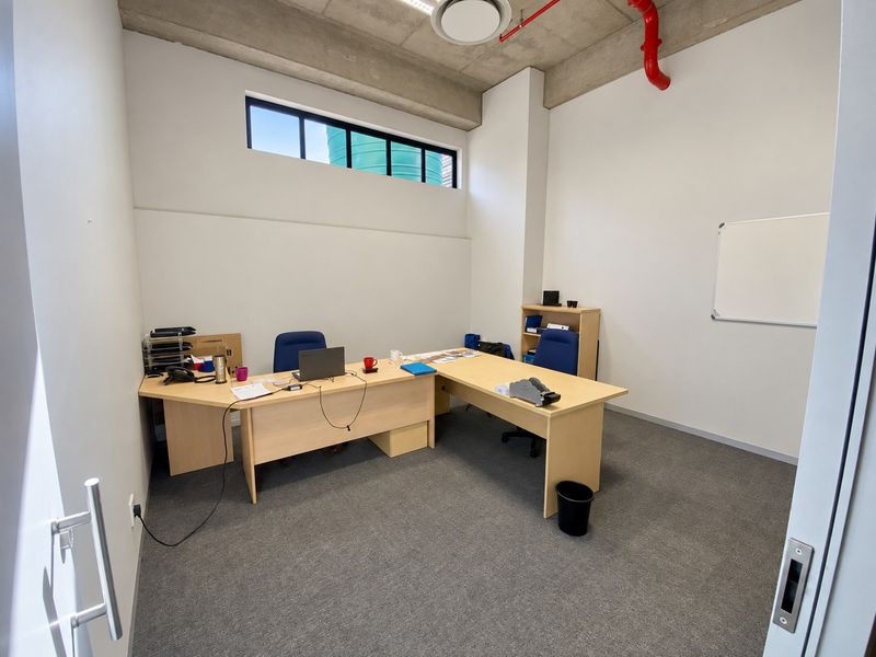 A functional office space featuring an exposed concrete ceiling with red fire sprinkler piping, carpeted flooring, and a high-level clerestory window for natural light.