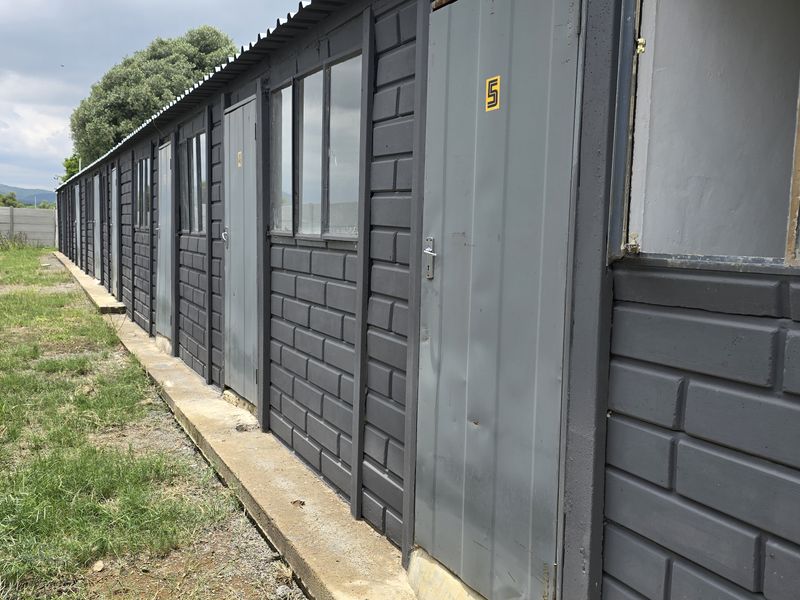 A row of modular commercial units featuring grey painted brick-style cladding, metal doors, and fixed windows, situated alongside a precast concrete boundary wall.