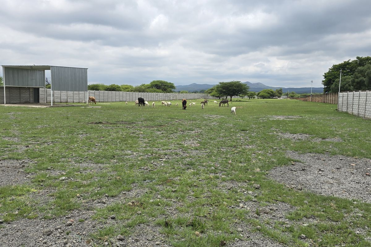 A large, open commercial vacant land plot enclosed by perimeter concrete block walls, featuring a metal storage structure and expansive grassy terrain.