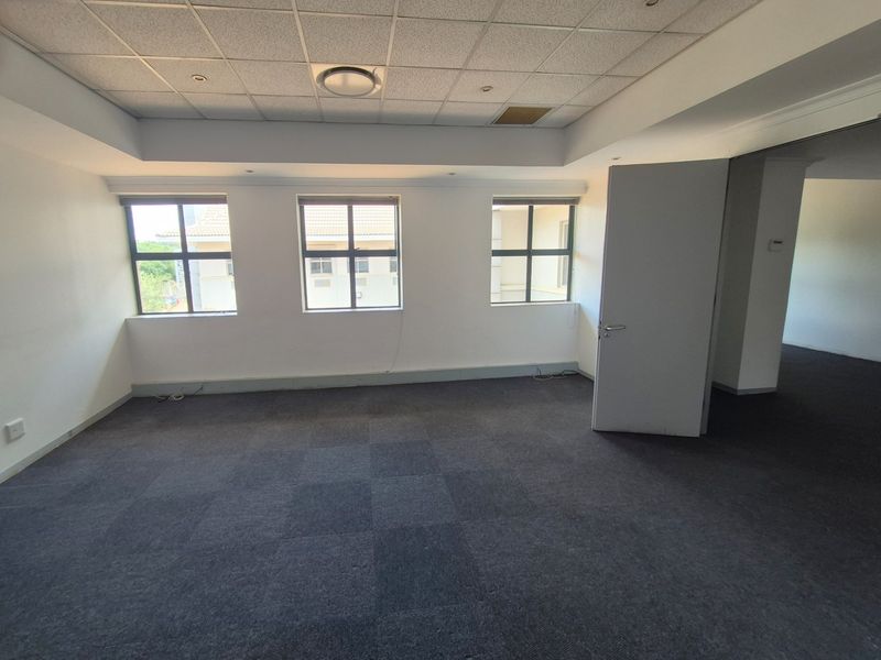 An empty, well-lit office space featuring carpeted flooring, a suspended ceiling with recessed lighting, and large windows providing natural light.