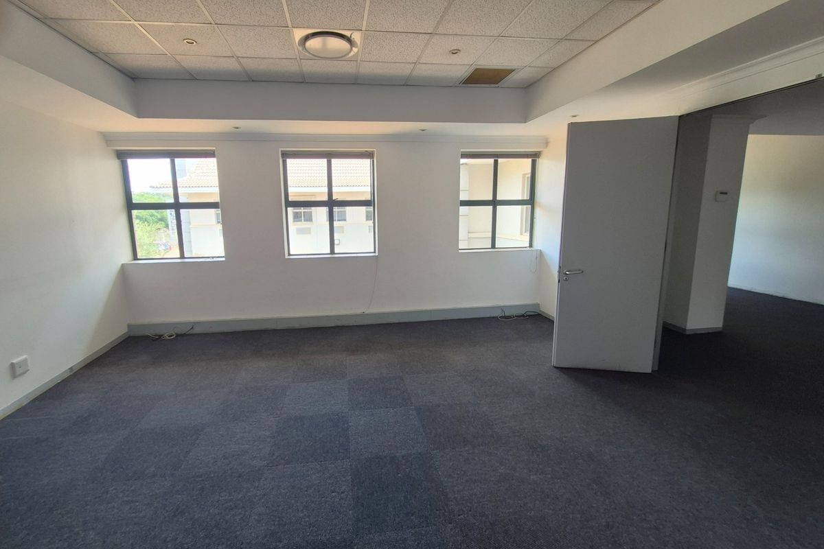 An empty, well-lit office space featuring carpeted flooring, a suspended ceiling with recessed lighting, and large windows providing natural light.