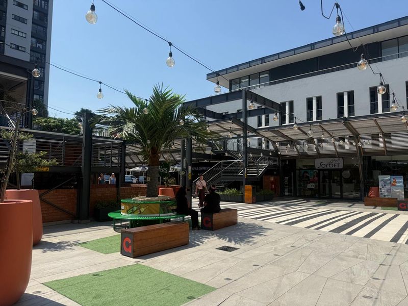 A vibrant outdoor retail courtyard featuring paved walkways, overhead string lighting, modern steel structures, and integrated seating areas within a commercial complex.