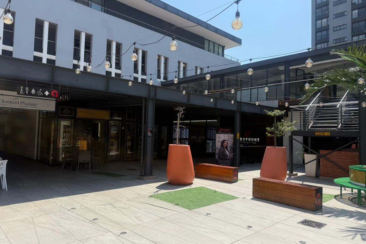 A modern outdoor retail courtyard featuring paved flooring, steel structural beams, overhead string lighting, and integrated commercial storefronts.