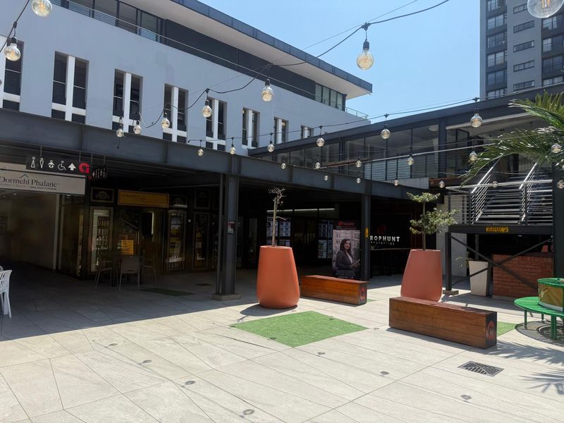 A modern outdoor retail courtyard featuring paved flooring, steel structural beams, overhead string lighting, and integrated commercial storefronts.