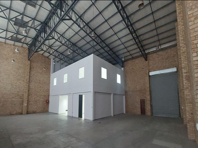 Spacious industrial warehouse featuring high ceilings with exposed steel trusses, concrete flooring, exposed brick walls, and a built-in mezzanine office structure.