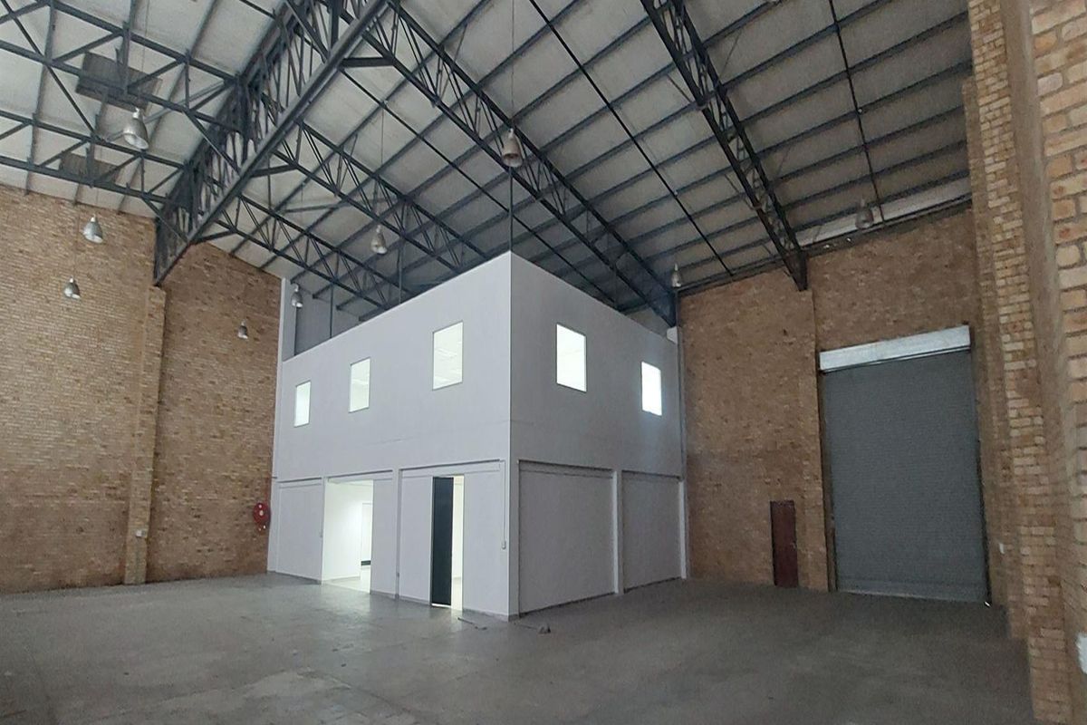 Spacious industrial warehouse featuring high ceilings with exposed steel trusses, concrete flooring, exposed brick walls, and a built-in mezzanine office structure.