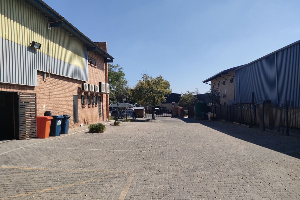 An industrial exterior view showing a paved yard area between two warehouse buildings with brick and metal cladding finishes.