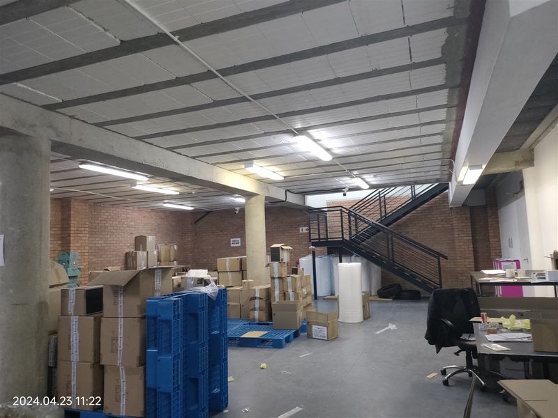 Industrial warehouse space featuring exposed brick walls, concrete ceiling, and a metal staircase leading to an upper level.