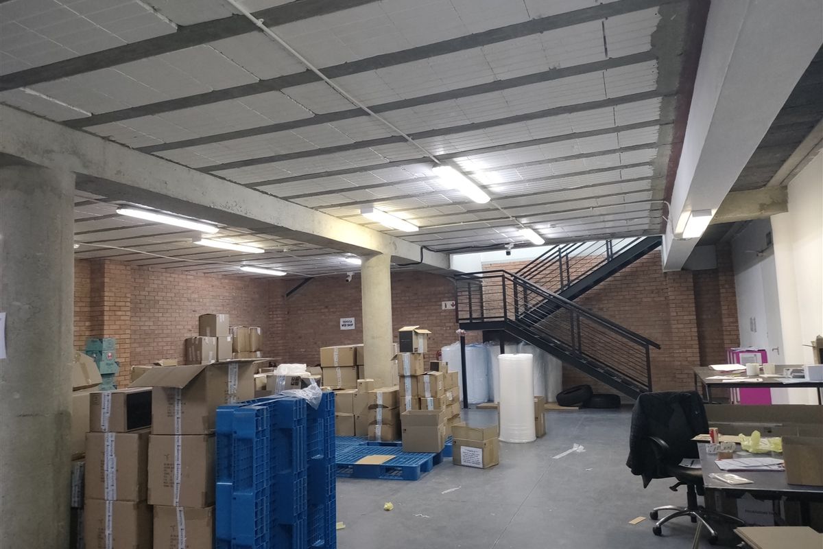 Industrial warehouse space featuring exposed brick walls, concrete ceiling, and a metal staircase leading to an upper level.