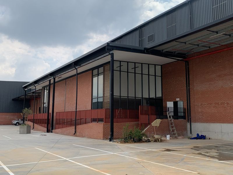 Modern industrial building exterior featuring red brick facade, large glass windows, metal roofing, and a concrete parking area with an accessible ramp.
