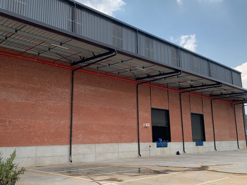 Industrial warehouse exterior featuring exposed brickwork, concrete plinth, roller shutter loading doors, and external fire suppression piping.