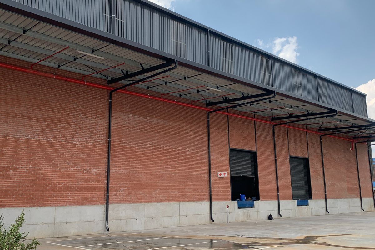 Industrial warehouse exterior featuring exposed brickwork, concrete plinth, roller shutter loading doors, and external fire suppression piping.