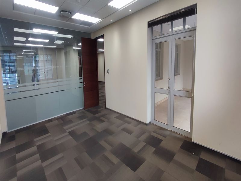A modern, empty office space featuring carpeted flooring, recessed ceiling lights, a glass partition wall, and a double glass door entrance.