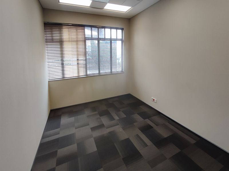A clean, empty office space featuring neutral walls, carpet tile flooring, recessed ceiling lighting, and large windows with fitted venetian blinds.