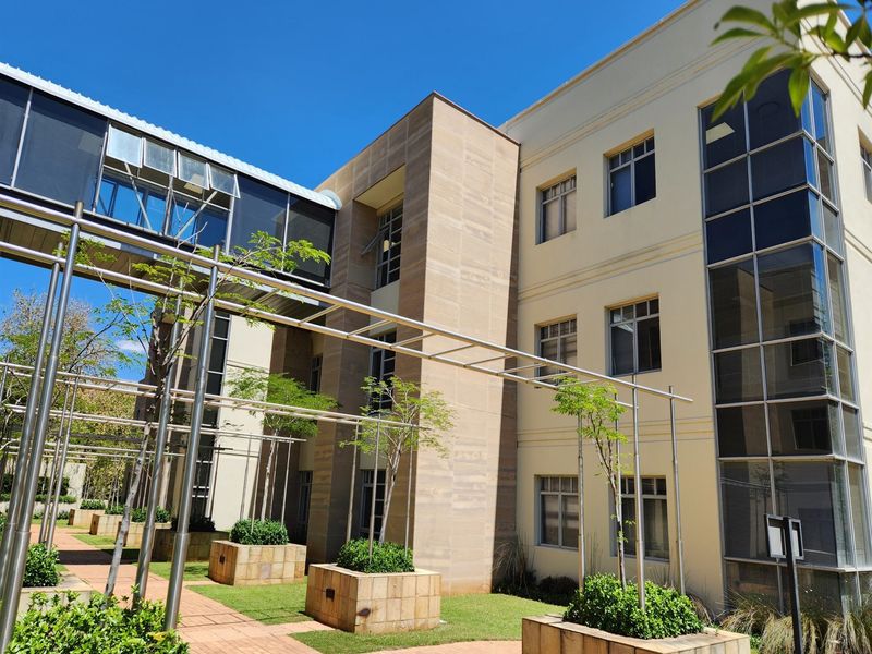 Modern office building exterior featuring a glass skywalk, stone-clad planters, and a paved courtyard area with metal pergola structures.
