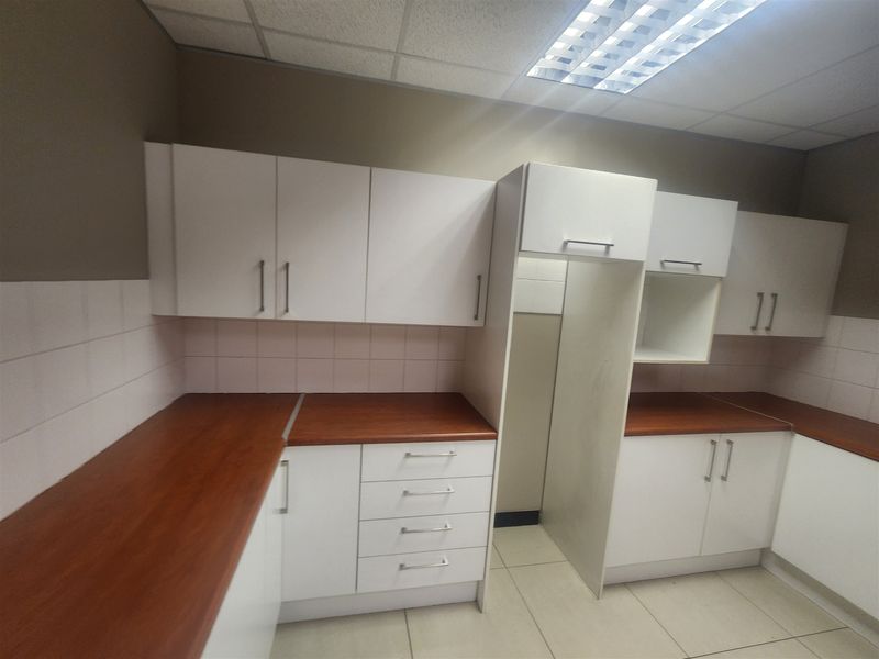 A clean, functional office kitchenette featuring white cabinetry, wood-look laminate countertops, tiled backsplash, and practical overhead fluorescent lighting.