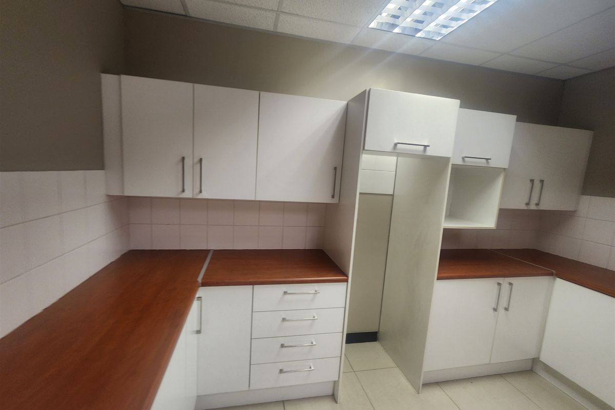 A clean, functional office kitchenette featuring white cabinetry, wood-look laminate countertops, tiled backsplash, and practical overhead fluorescent lighting.