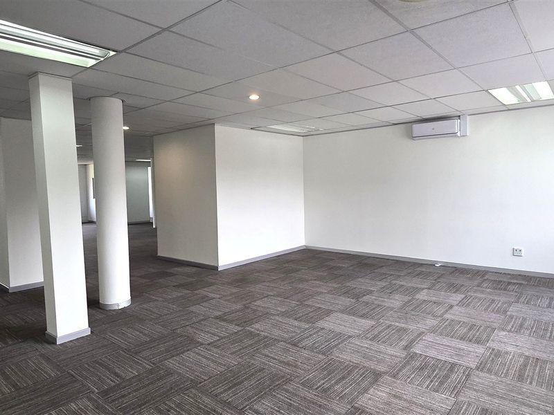Spacious, open-plan office area featuring carpet tiles, a suspended ceiling with recessed lighting, and a wall-mounted air conditioning unit.