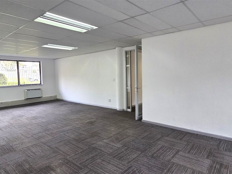 A spacious, empty office area featuring carpet tiles, a suspended ceiling with recessed lighting, and wall-mounted HVAC units.