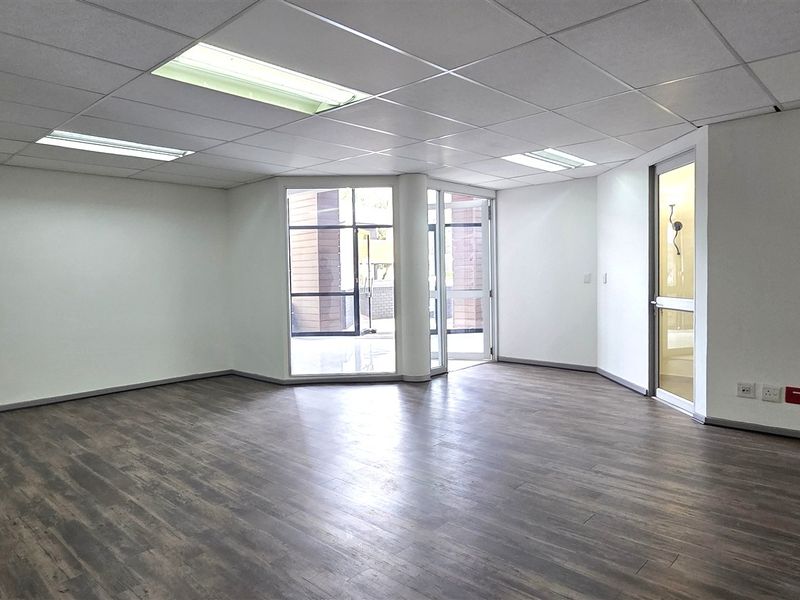 A bright, open-plan office space featuring modern laminate flooring, a suspended ceiling with recessed lighting, and a wall-mounted air conditioning unit.