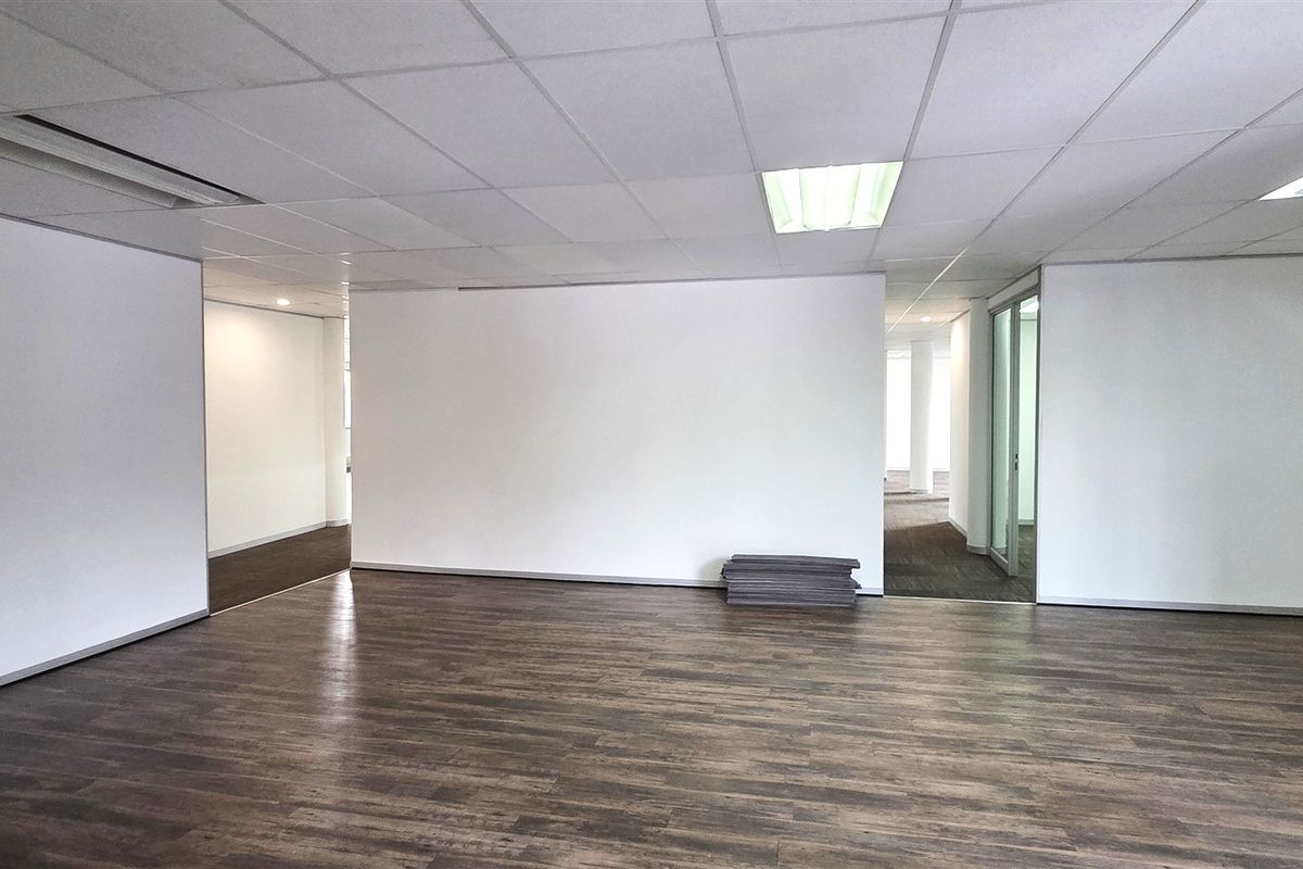A spacious, empty office area featuring a suspended ceiling with recessed lighting and dark wood-look laminate flooring, offering a clean and professional workspace.