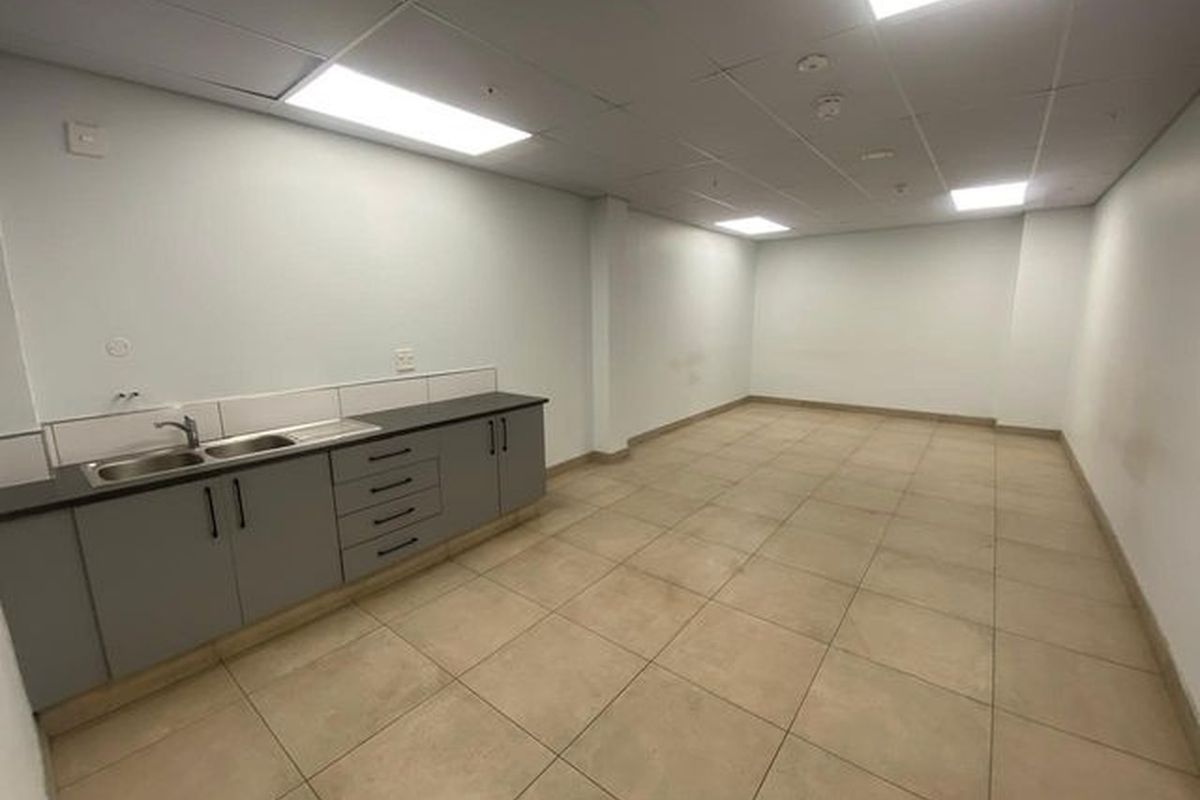 A clean, empty industrial kitchenette featuring grey built-in cabinetry, a stainless steel sink, tiled flooring, and recessed lighting in a suspended ceiling.